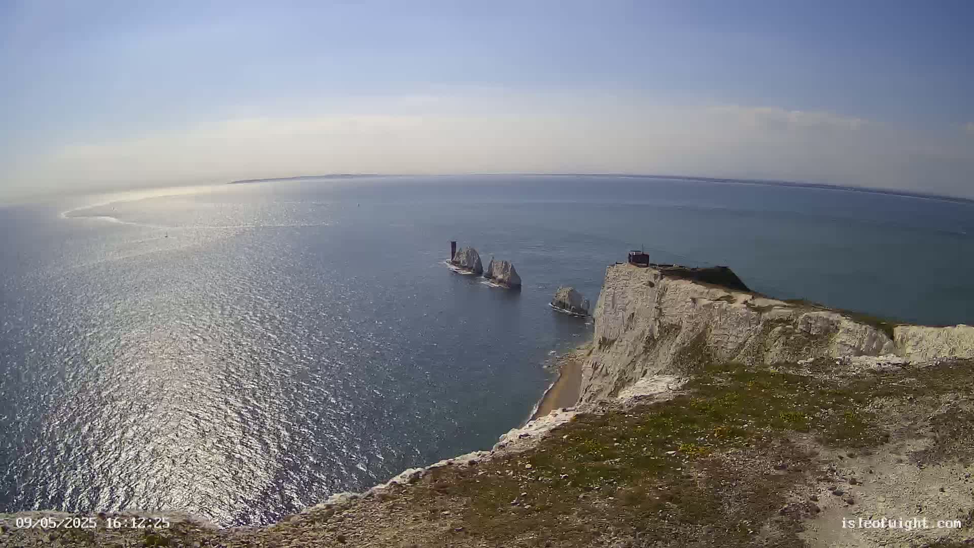 A high-angle view shows a sunny day with a calm sea, white cliffs, and several small, white rock formations extending into the water.