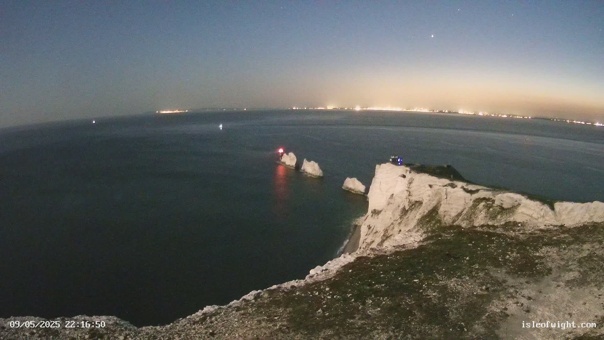 A nighttime coastal scene shows white cliffs overlooking dark water, with three stacks of rocks and a distant shoreline visible under a clear, dark sky.