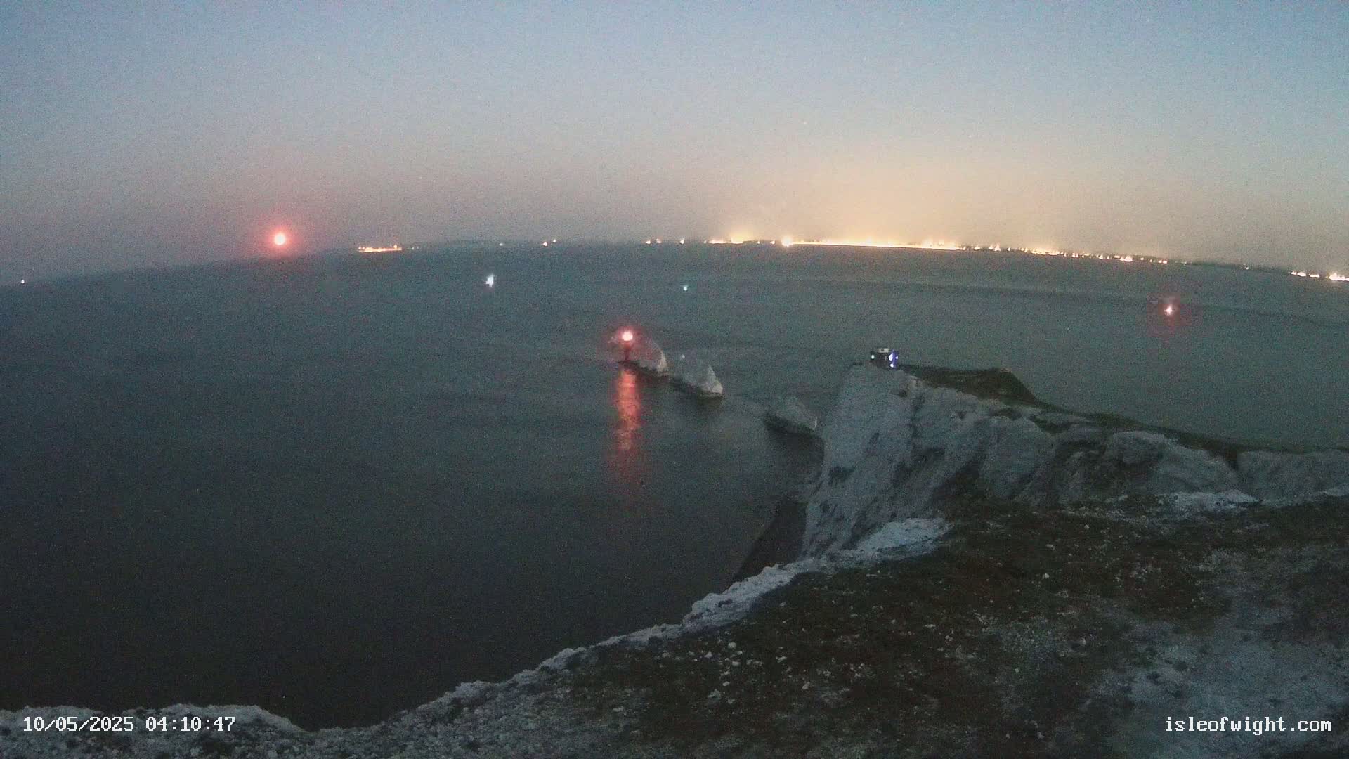A dark, twilight scene shows a rocky cliff overlooking a calm sea with a lit lighthouse and distant city lights on the horizon.