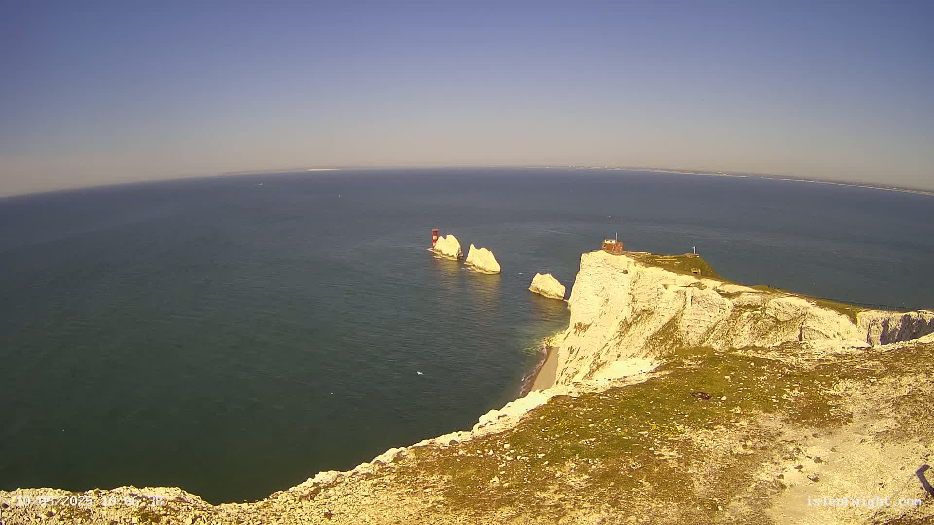 A high-angle view on a sunny day shows white cliffs overlooking a calm sea where three stacks of rock and a small building stand out.
