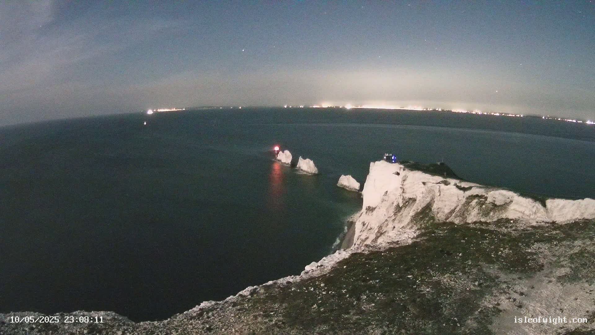 A nighttime view from a clifftop overlooking a dark sea with white rocks and a distant, faintly lit coastline under a clear, starry sky.