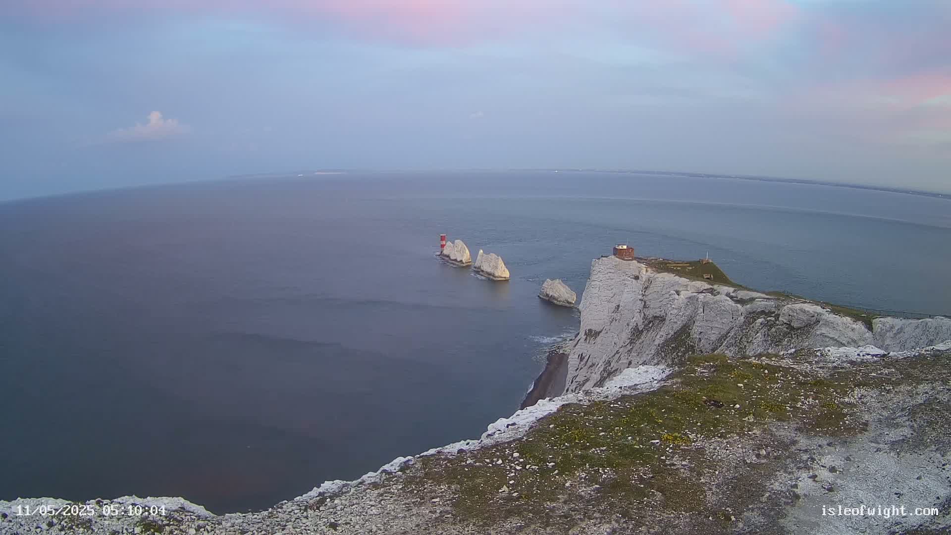 White cliffs overlook a calm sea with three white rock formations and a small red lighthouse in the distance under a pale, cloudy sky.