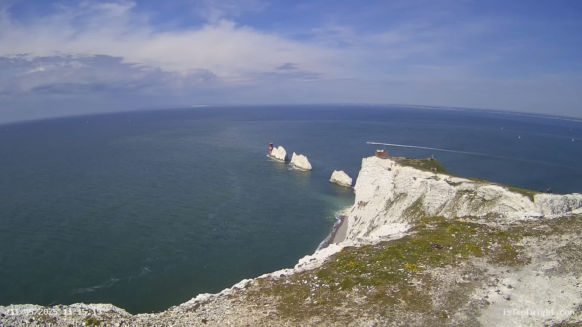 White cliffs overlook a calm, blue sea with several small, white rock formations and a small building on one of the cliffs, under a partly cloudy sky.