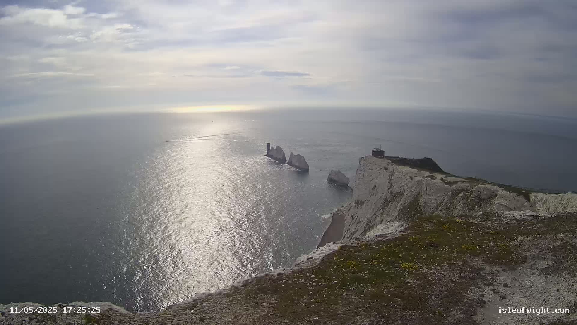 A high-angle view shows white cliffs overlooking a calm, sunlit sea with several stacks of rock visible in the distance under a partly cloudy sky.