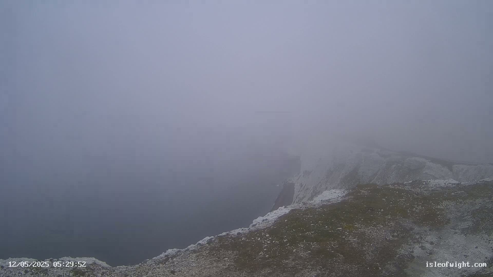 A foggy view from a clifftop shows a steep drop to a barely visible expanse of water below.