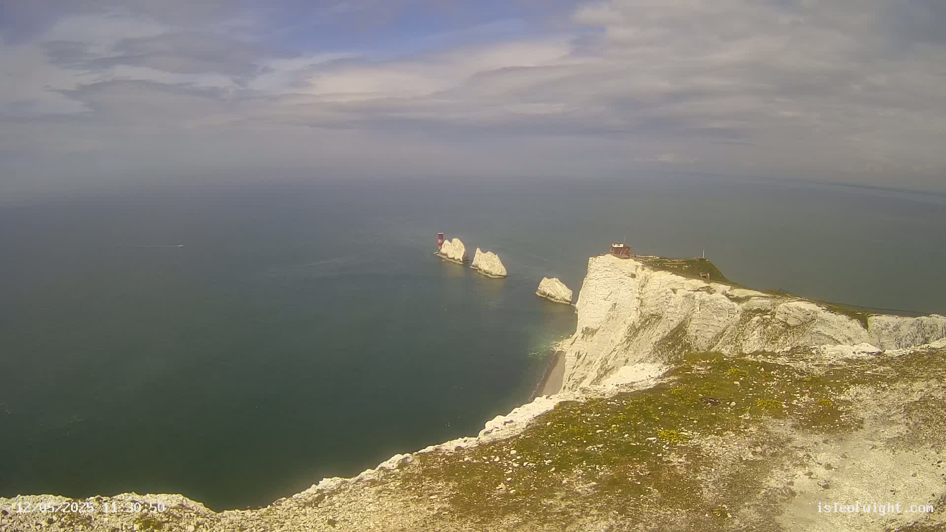 White cliffs meet a calm, grey-blue sea, with three smaller chalk stacks and a small building visible offshore under a partly cloudy sky.