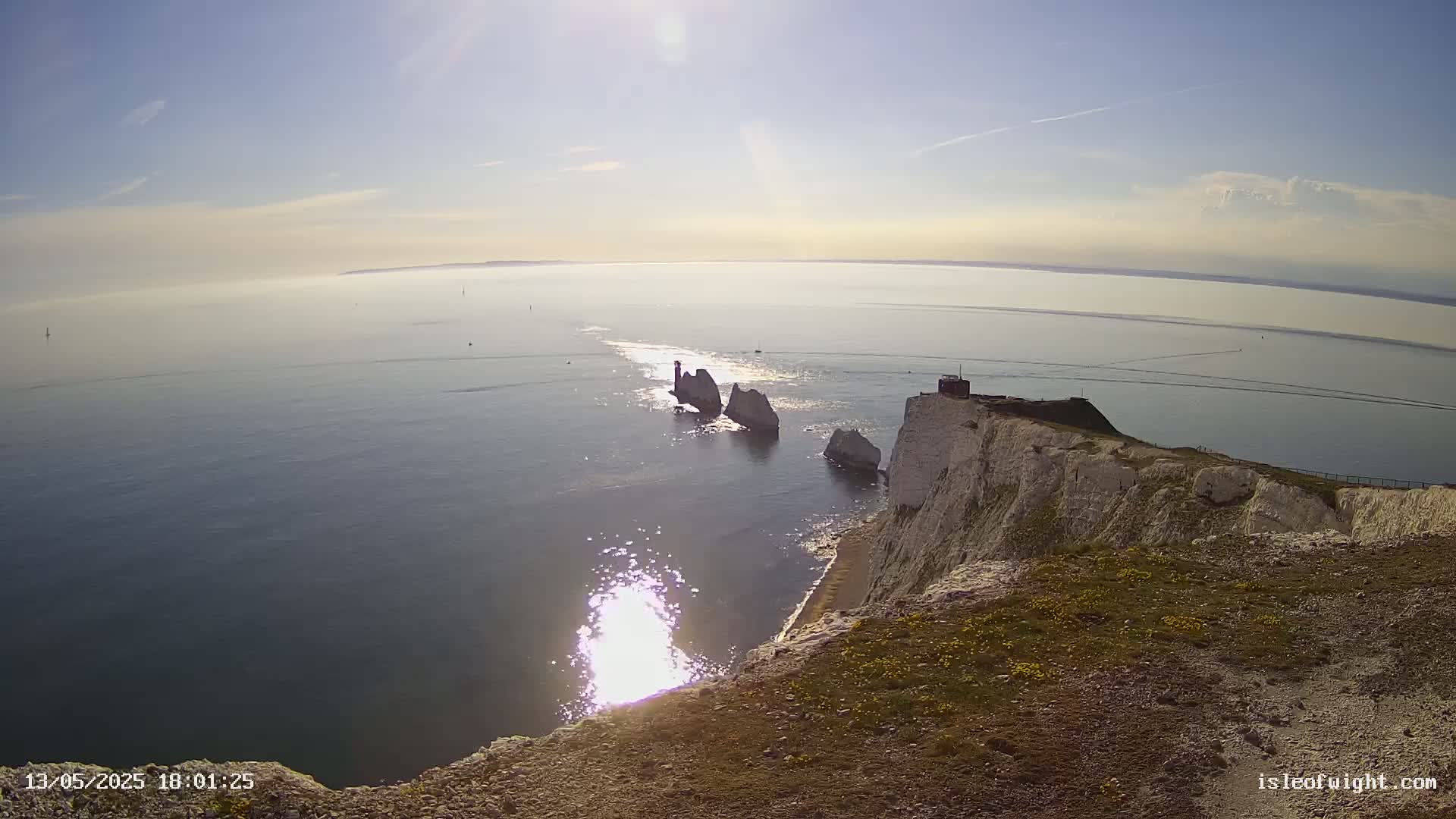 A high-angle view shows white cliffs overlooking a calm sea, where several rocky outcrops are visible in the distance under a bright, sunny sky.