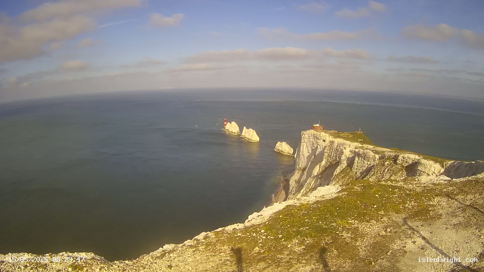 A high-angle view shows a partly cloudy sky over a calm sea, white cliffs, and three stacks of rock with a small building on one.
