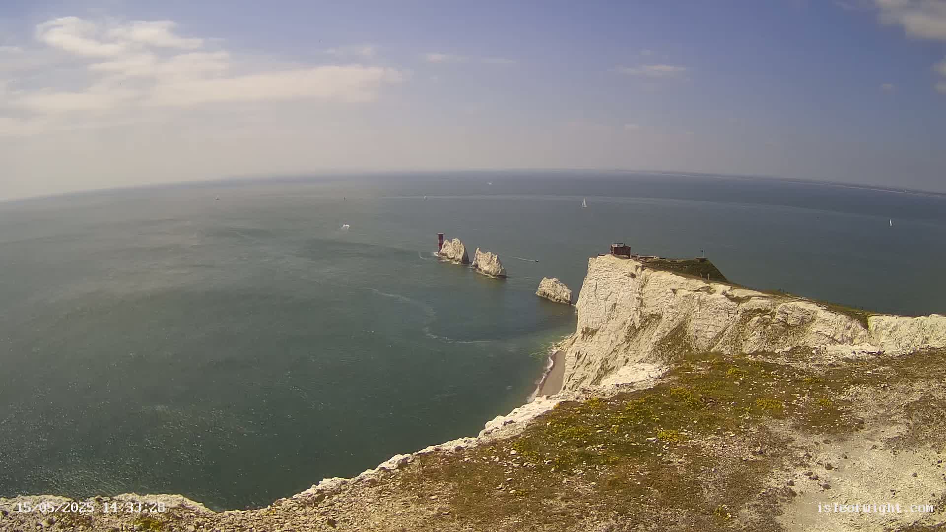 White cliffs overlook a calm sea with several small islands and a lighthouse visible in the distance under a partly cloudy sky.