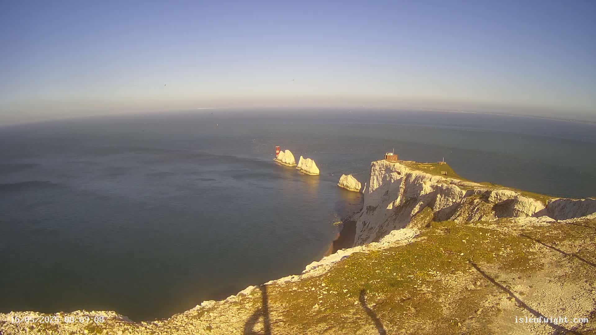 White cliffs overlooking a calm sea with three smaller, distant white rock formations, one of which has a small red and white structure on top.