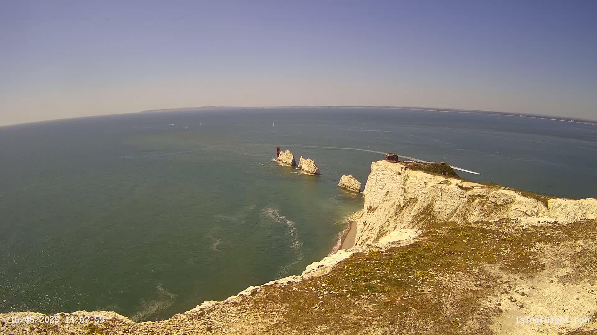 White cliffs overlooking a calm, green-blue sea with several stacks of rock formations and a small building visible in the distance under a clear, sunny sky.
