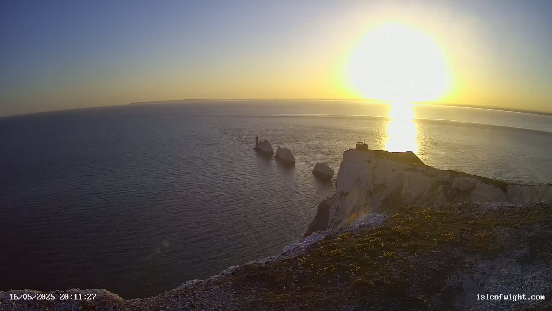 A bright sun sets over a calm sea, illuminating white cliffs and several stacks of rock jutting out from the water.