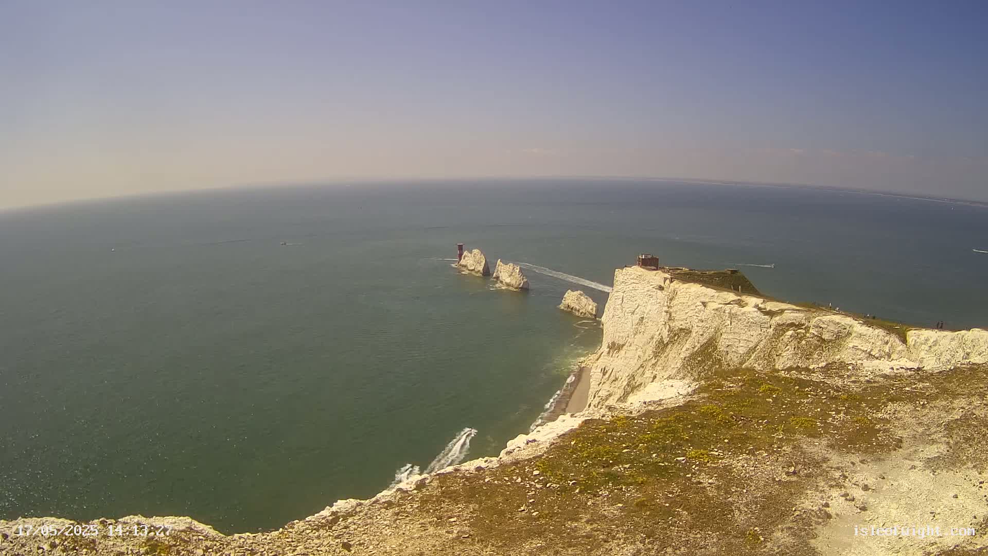 A high-angle view on a sunny day shows a white cliff overlooking the ocean, with several smaller white rock formations and a small building visible in the distance.