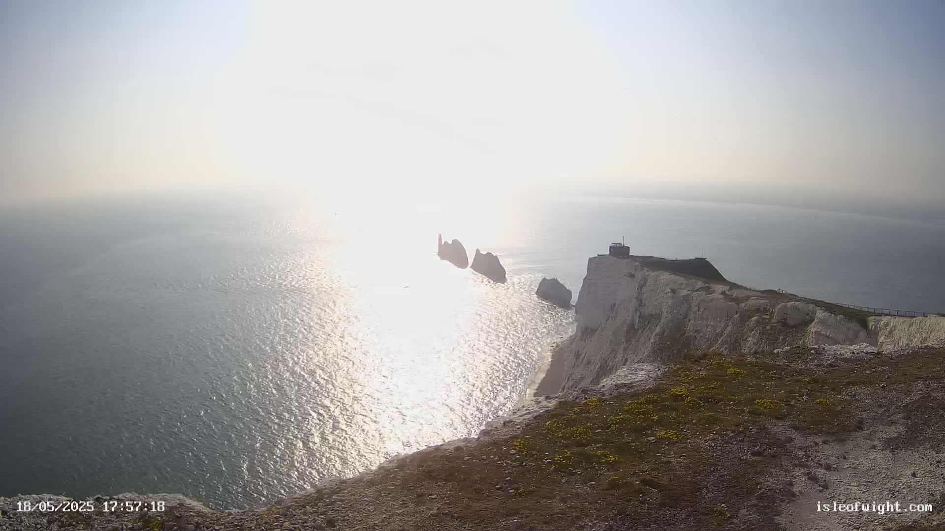 White cliffs overlook a sparkling sea under a bright, hazy sun, with several small islands visible in the distance.