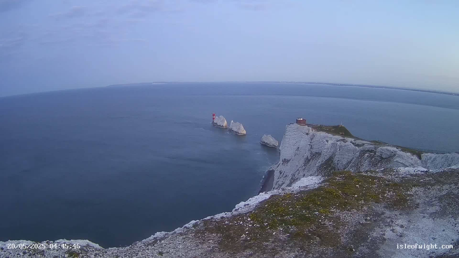 White cliffs overlook a calm sea where several white rock formations and a red lighthouse are visible in the distance under a clear sky.
