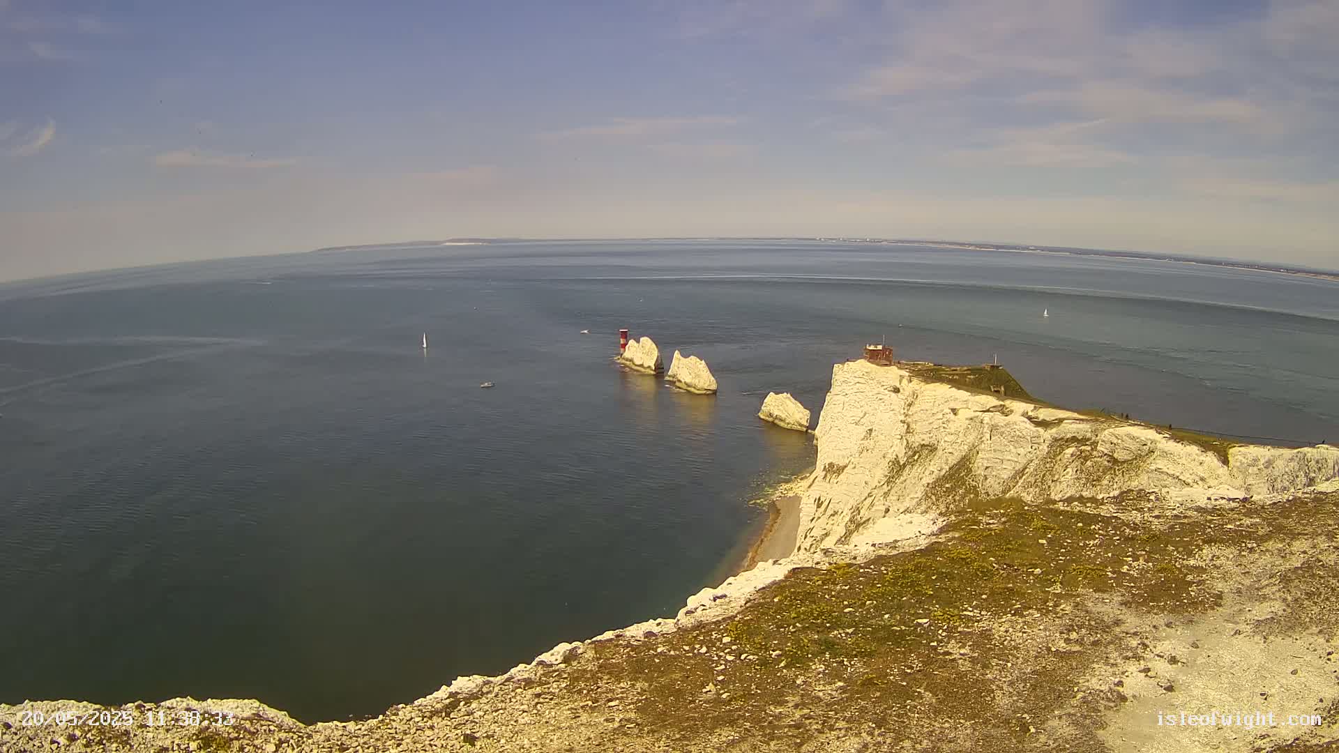 A high-angle view shows white cliffs overlooking a calm sea, where three stacks of rock rise from the water, one topped with a small structure.