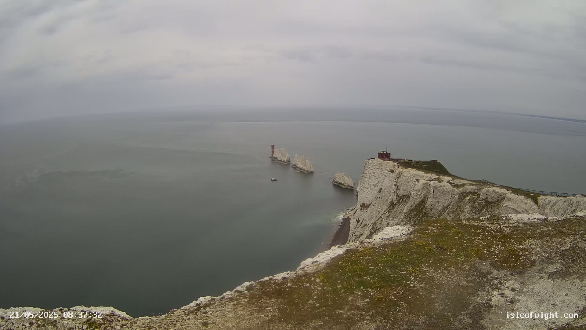 A high-angle view shows white cliffs overlooking a calm, gray sea with several small islands and a small structure on one of the cliffs under an overcast sky.