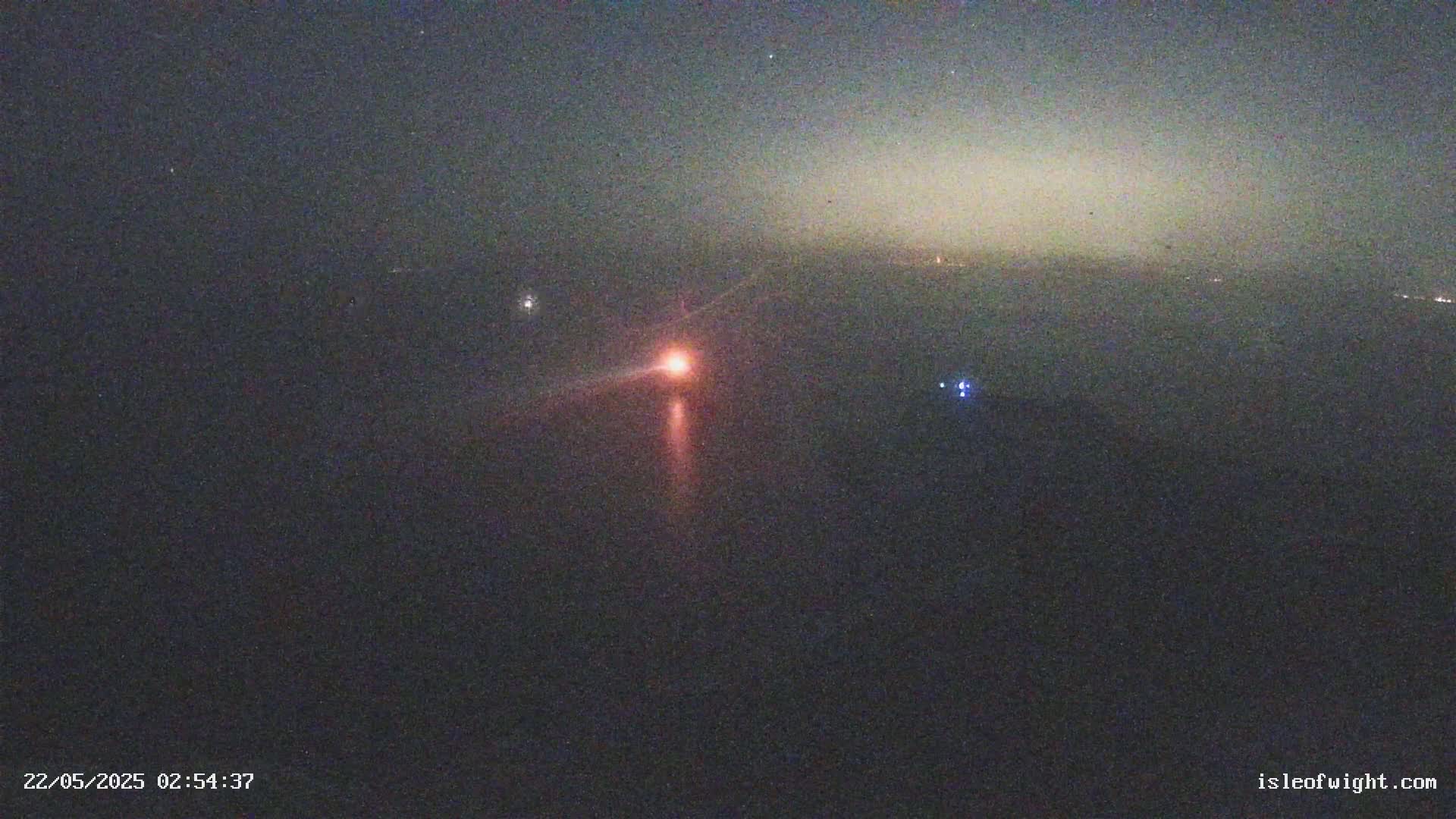 A nighttime aerial view shows a single bright orange light on the horizon with a faint reddish glow emanating from it, under a dark, mostly clear sky.