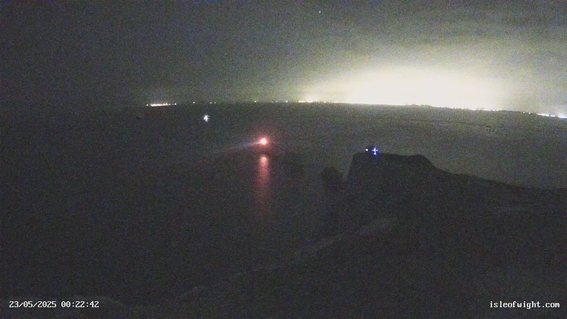 A nighttime long shot shows a distant line of city lights and a single vehicle with red taillights driving on a dark road.