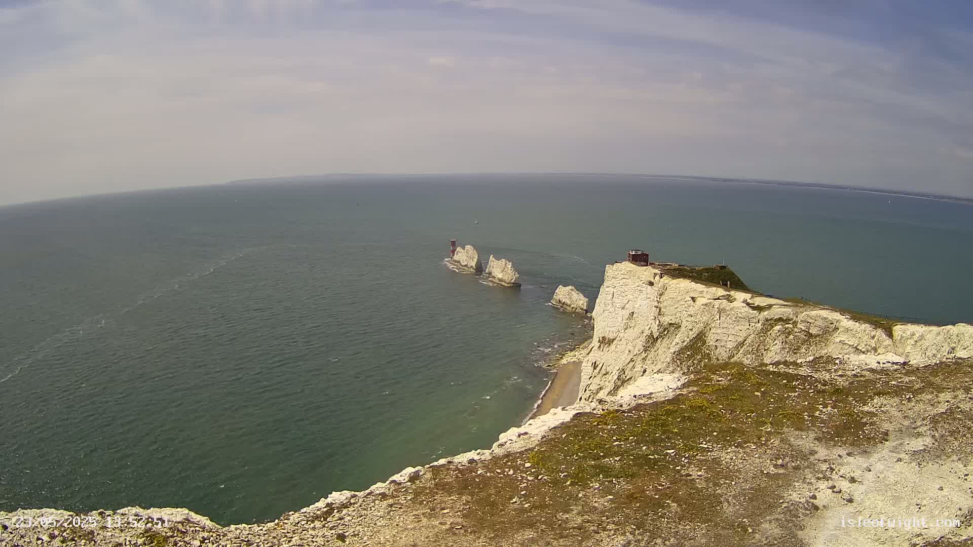 A high-angle view shows a rocky, white cliff overlooking a calm sea with several smaller rock formations extending out into the water on a partly sunny day.
