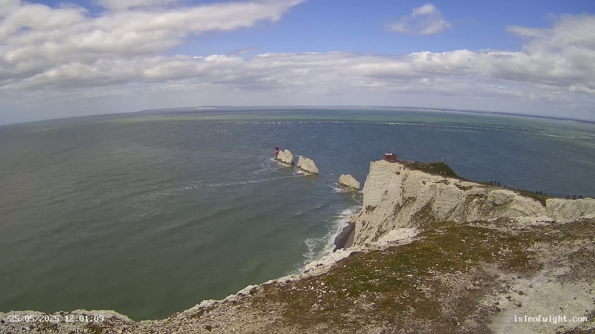 White cliffs overlook a choppy sea with three stacks of rock and a small building in the distance under a partly cloudy sky.