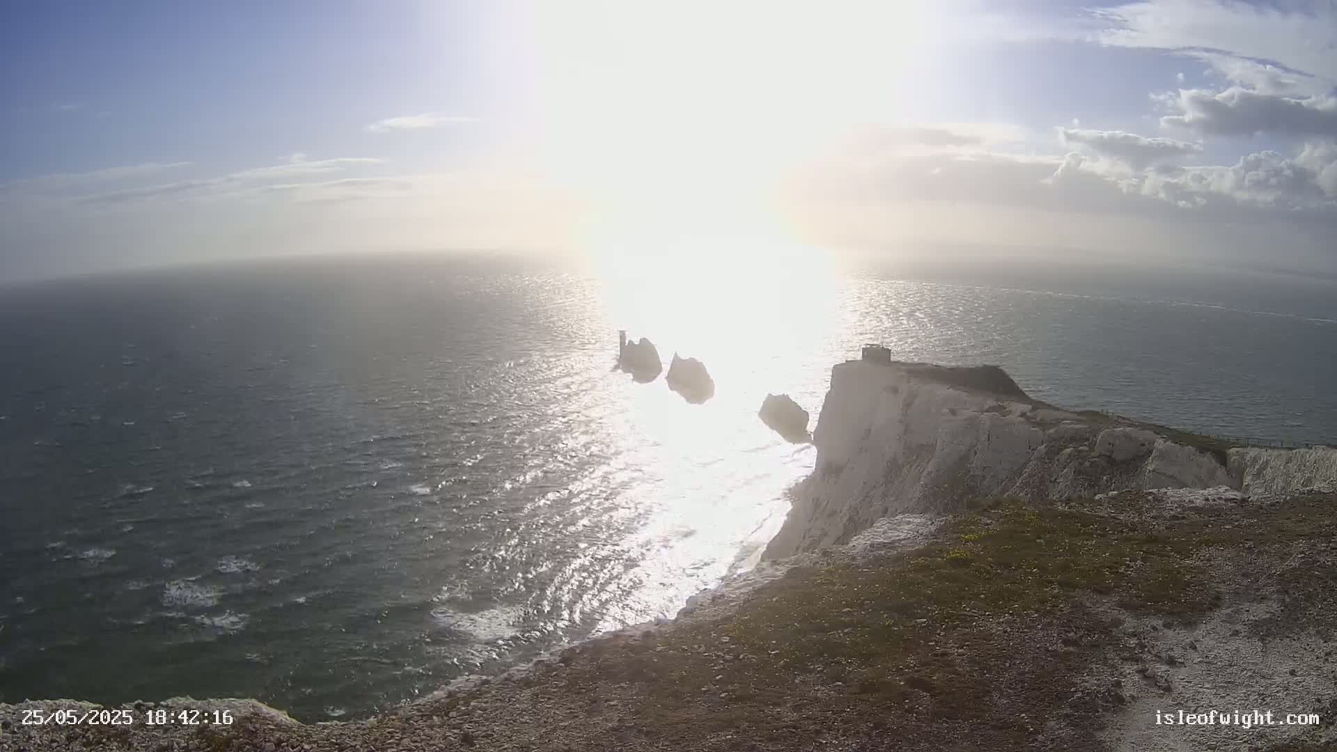 White cliffs overlook a wavy sea with several small islands in the distance, under a bright, partly cloudy sky.