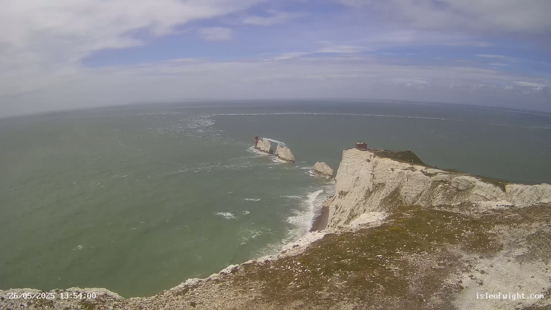 A high-angle view shows white cliffs meeting a choppy green sea, with a small island and lighthouse visible in the distance under a partly cloudy sky.