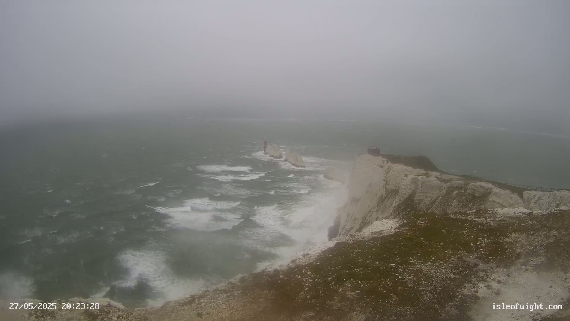 White cliffs meet a rough, gray sea under a foggy sky, with waves crashing against the rocks and a small structure visible on the clifftop.