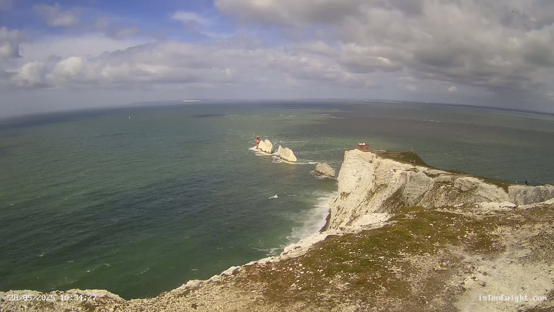 White cliffs overlooking a green sea with a few small islands and a lighthouse in the distance under a partly cloudy sky.
