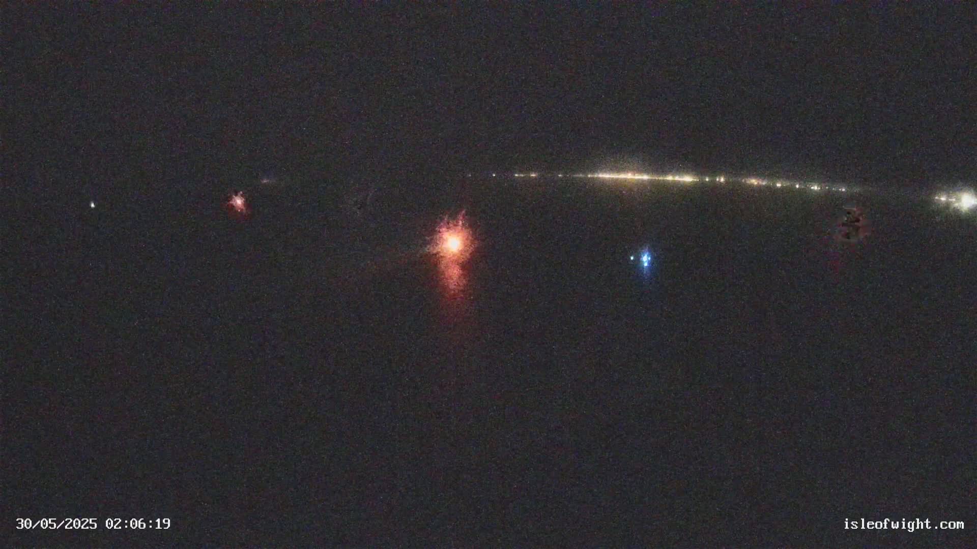 A nighttime, dark, low-light image shows a distant line of lights, possibly a town, with several smaller, brighter lights closer to the viewer, one appearing reddish-orange.