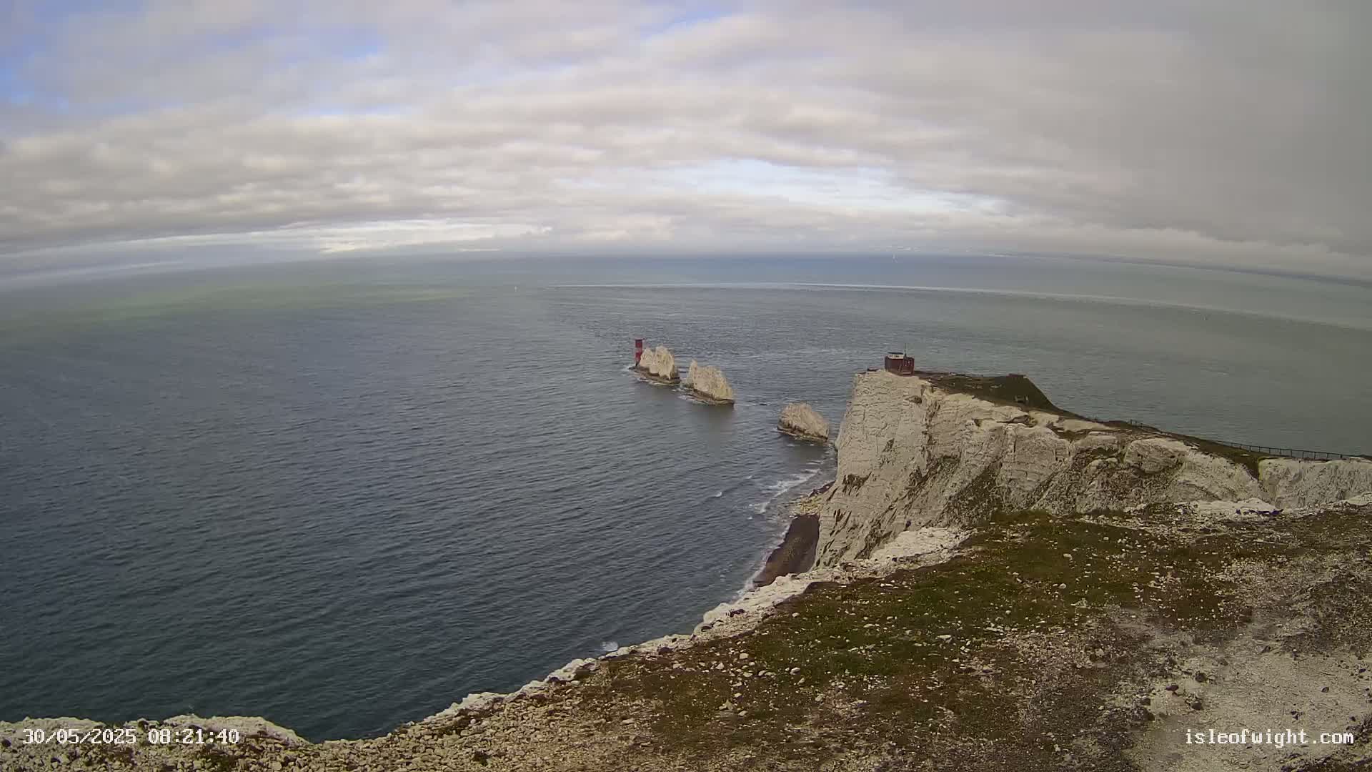 A high-angle view shows white cliffs overlooking a dark-blue sea, with a small group of rock formations and a small structure visible in the distance under a cloudy sky.