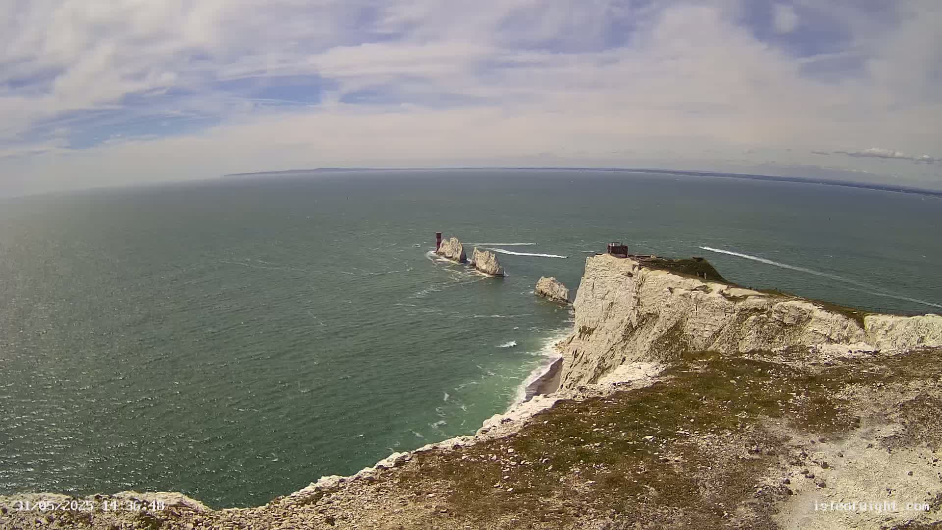 White cliffs overlooking a green ocean with several stacks of rock and a few boats visible in the distance under a partly cloudy sky.