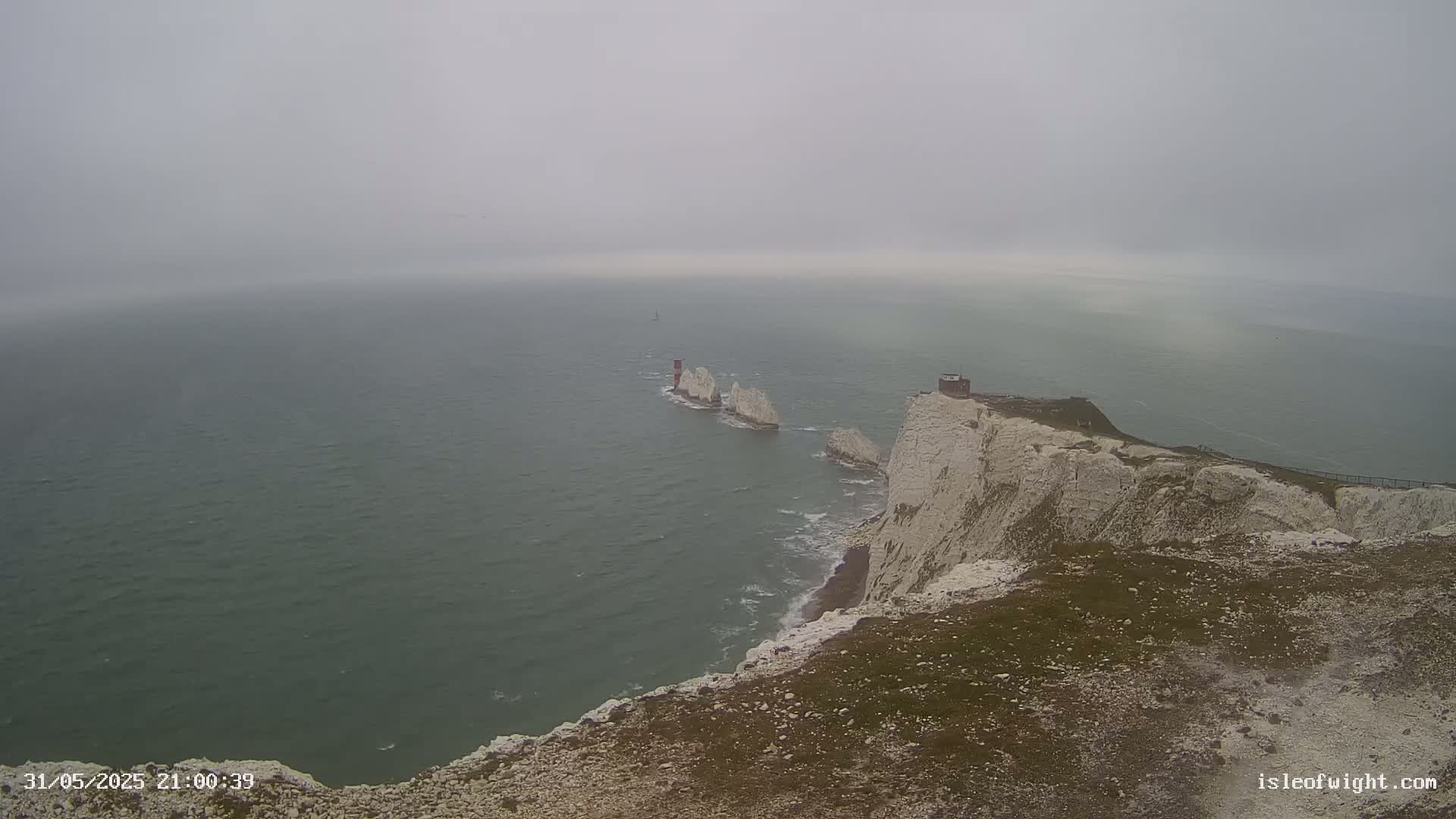 A foggy view of a white cliff overlooking a body of water with a line of stacks and a small building visible in the distance.