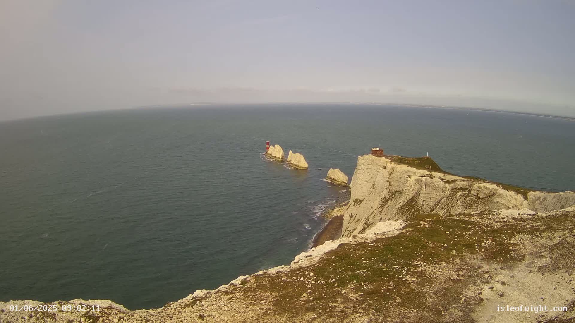 A hazy, light-colored sky overlooks a rocky, white cliff overlooking a dark-blue ocean, where three small white rock formations with a small red structure are visible in the water.