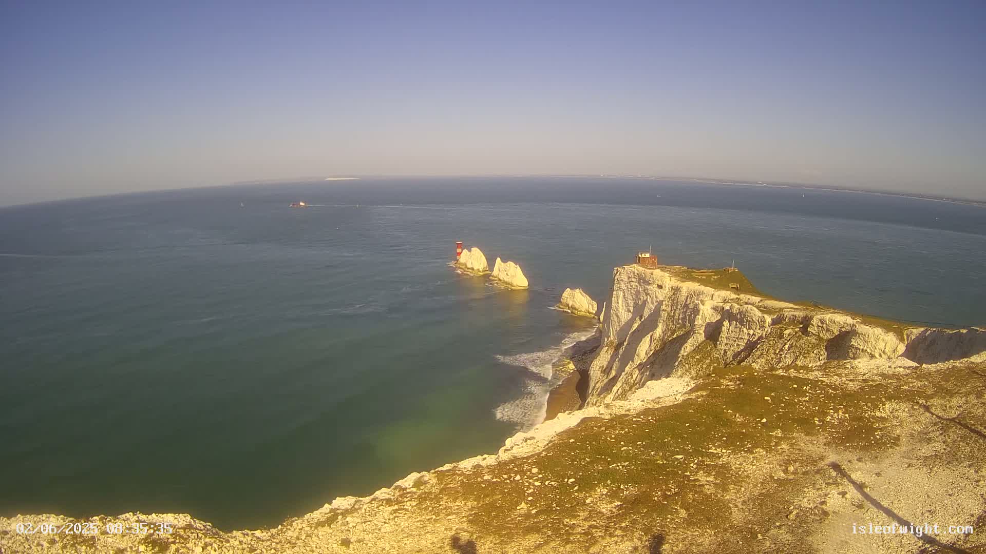 A high-angle view shows white cliffs overlooking a calm, greenish-blue ocean, with two small islands and a lighthouse visible in the distance under a clear blue sky.