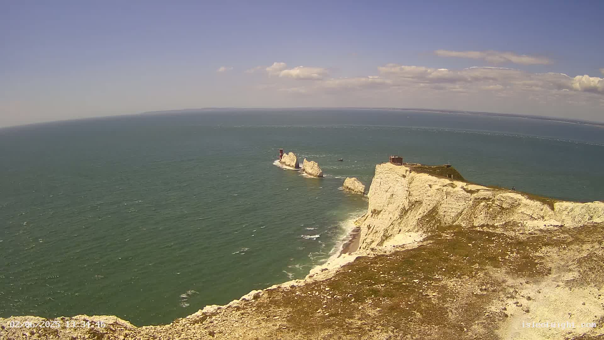White cliffs overlook a body of water, with three rock formations and a small structure visible in the distance under a mostly sunny sky with a few clouds.