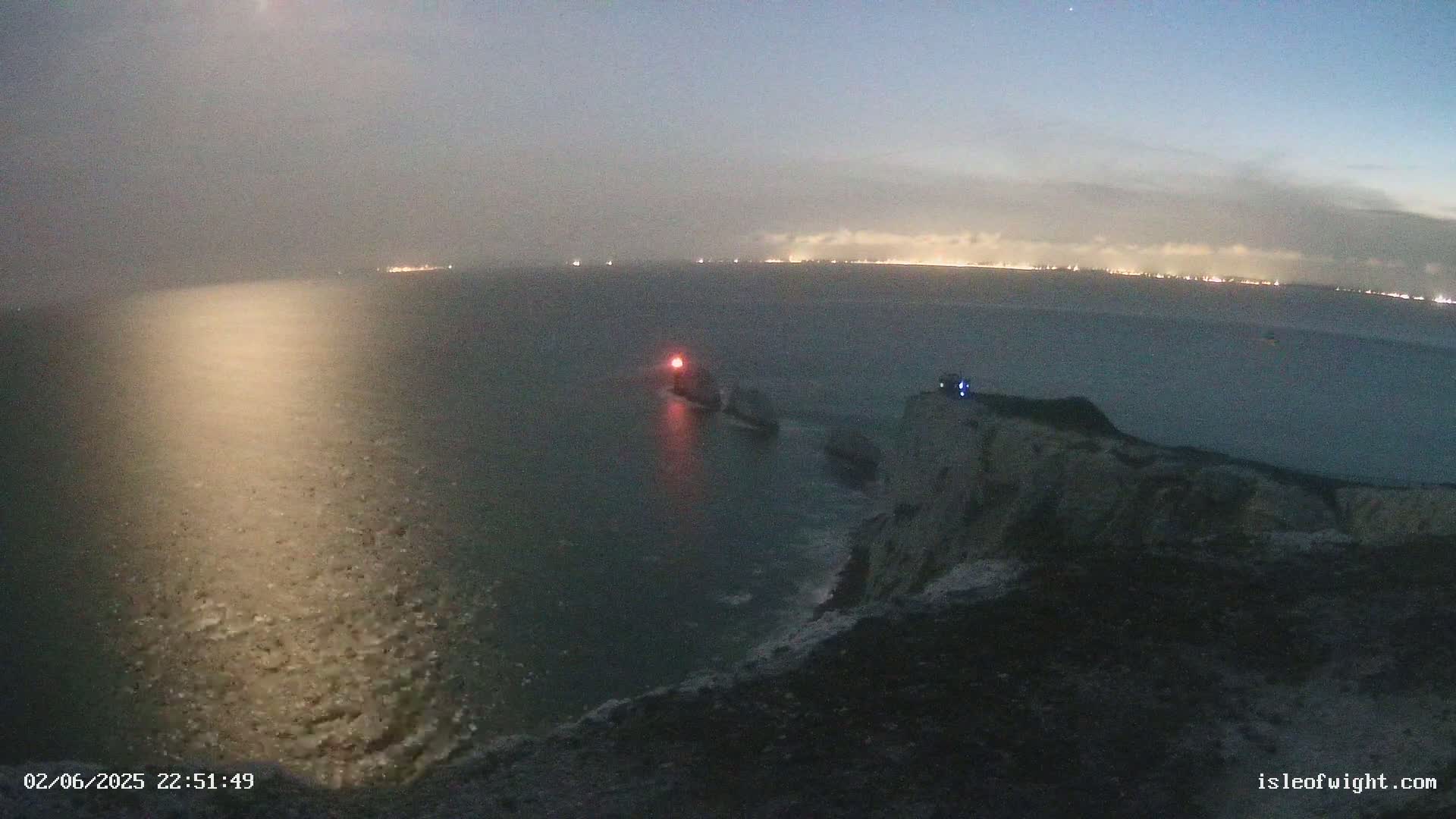 A dark, foggy night scene shows a body of water with a distant, faintly lit coastline, and a rocky cliff with a light visible on a structure near the water's edge.