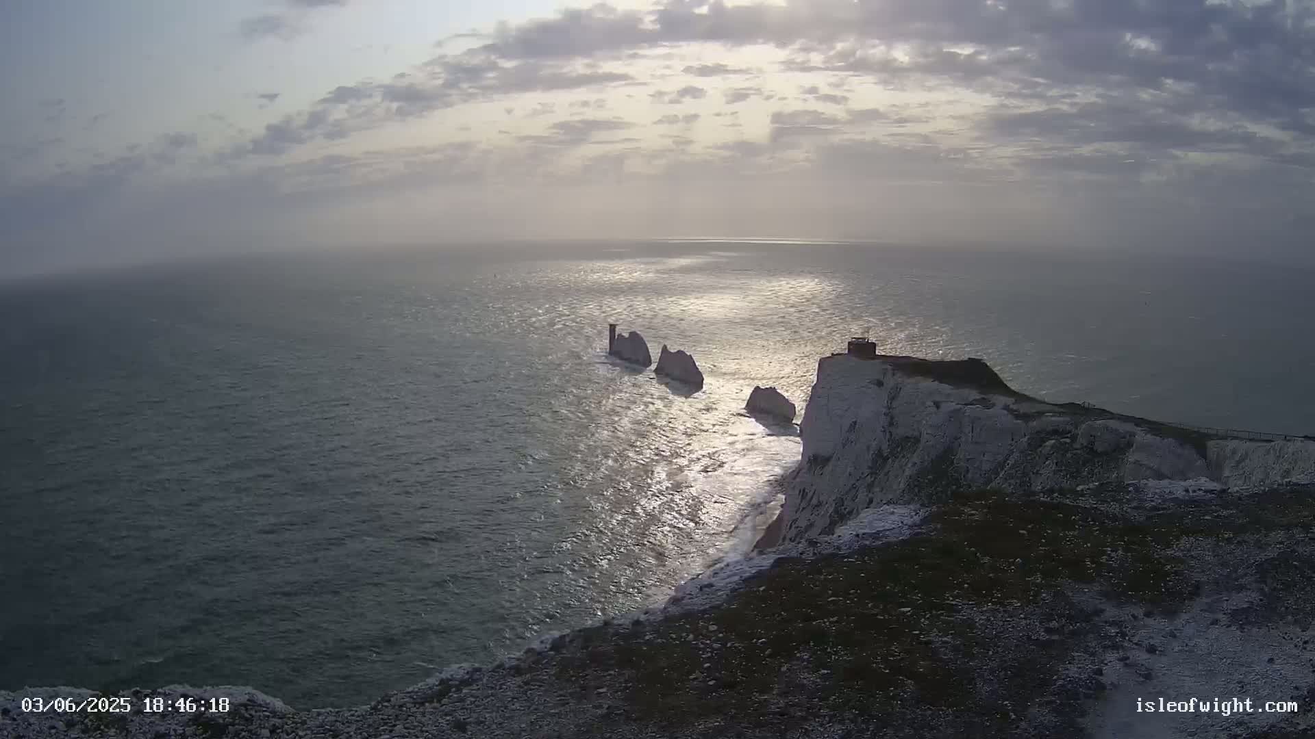 A high-angle view shows a rocky, white cliff overlooking a dark-blue sea where several stacks of rock are visible in the distance, under a mostly cloudy sky.
