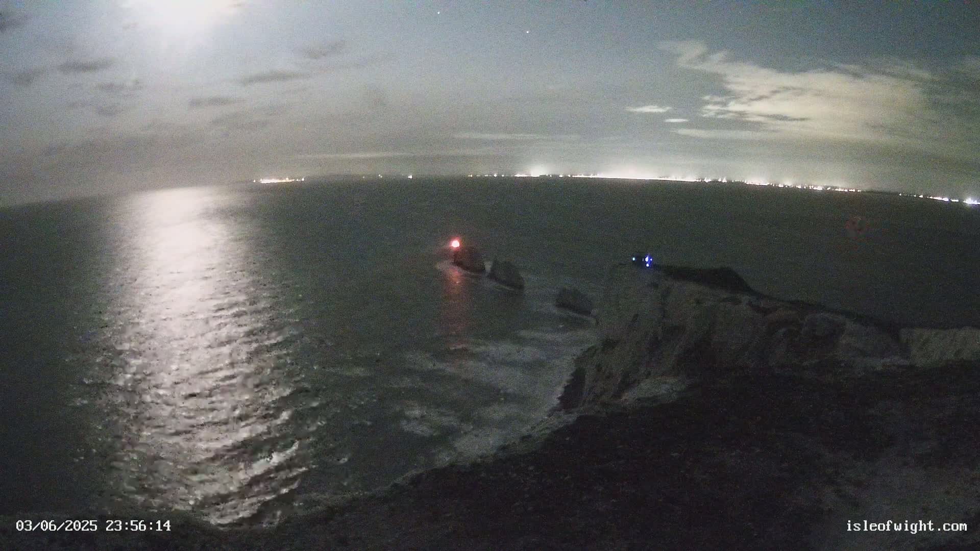 A nighttime coastal scene under a partly cloudy sky shows a moonlit sea with dark rocky land in the foreground, featuring a small light near some rocks in the water.