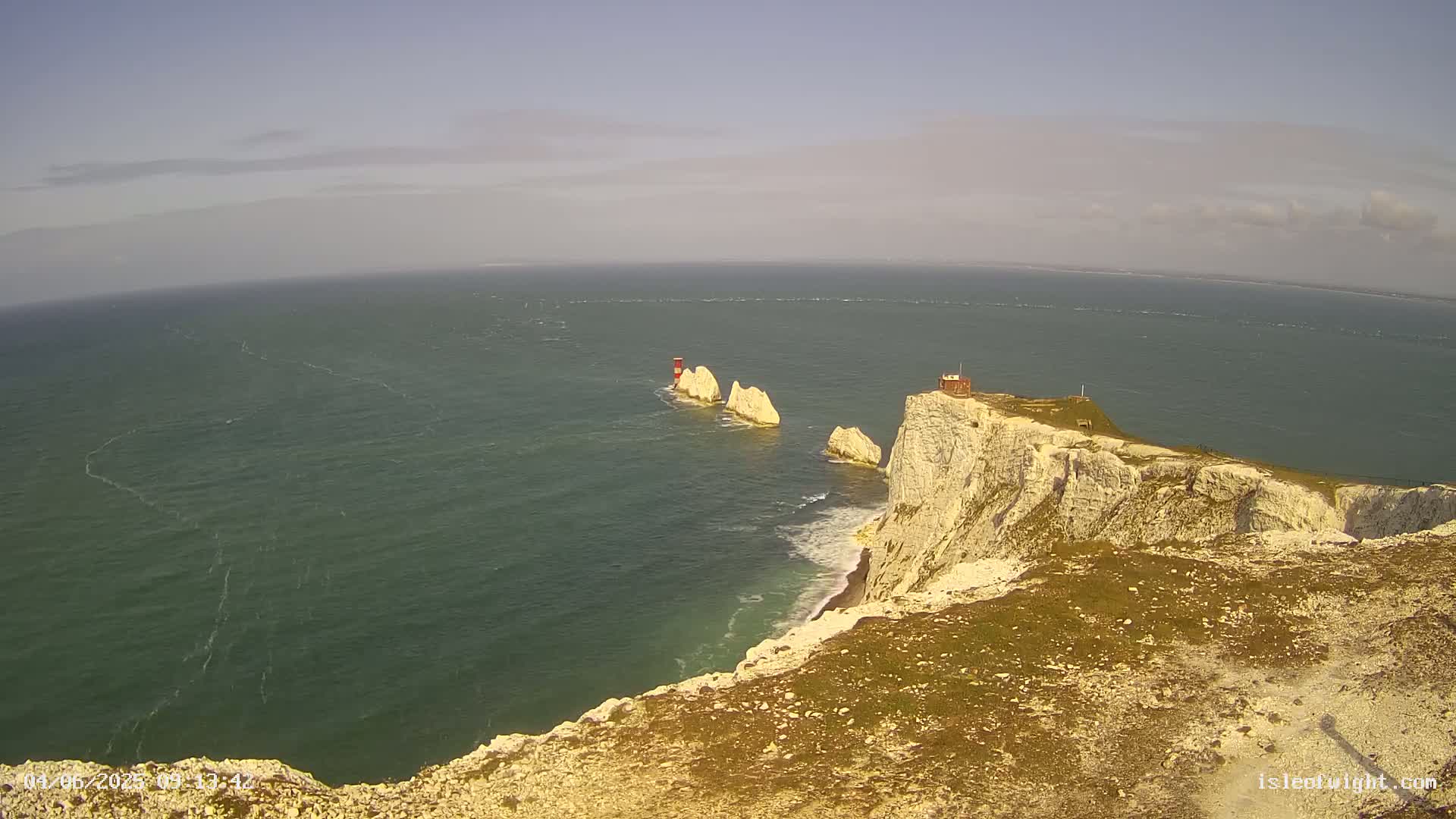 White cliffs meet a calm, greenish-blue sea where several stacks with a lighthouse are visible offshore.