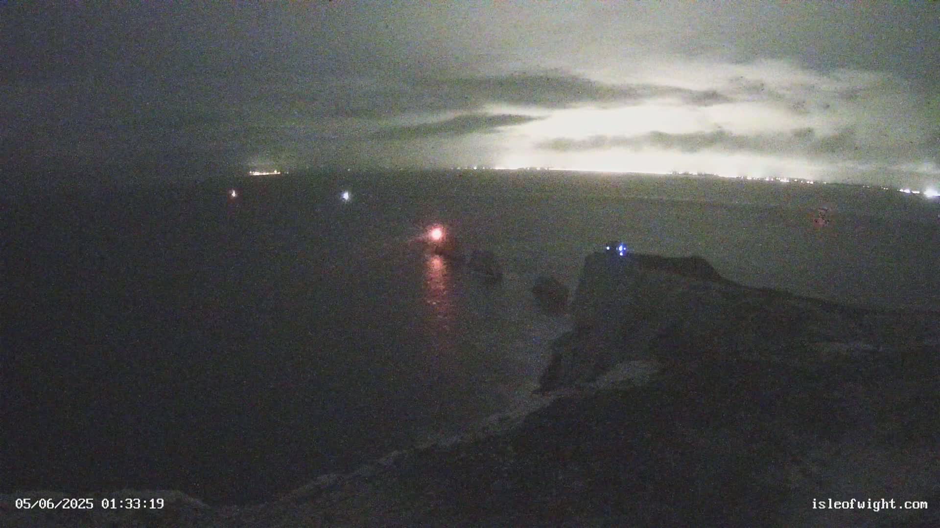 A nighttime coastal scene shows a dark body of water with a few faint lights visible in the distance under a cloudy sky.