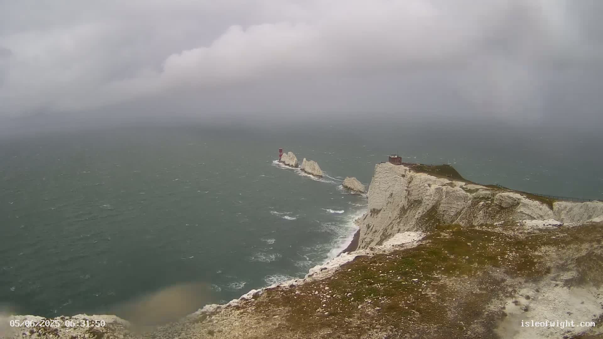 White cliffs overlook a choppy sea with a distant group of smaller rock formations and a lighthouse under an overcast sky.