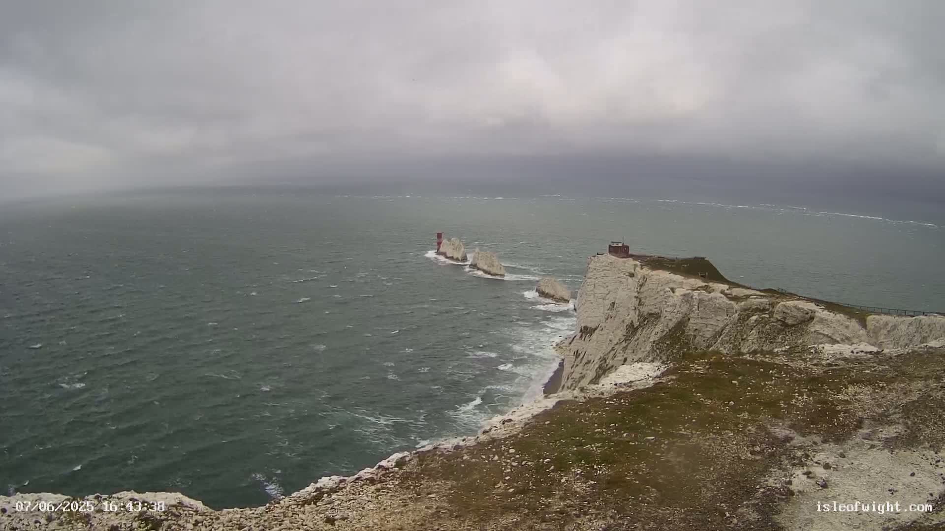 White cliffs overlook a choppy sea with several small rocky islands and a lighthouse under a cloudy sky.