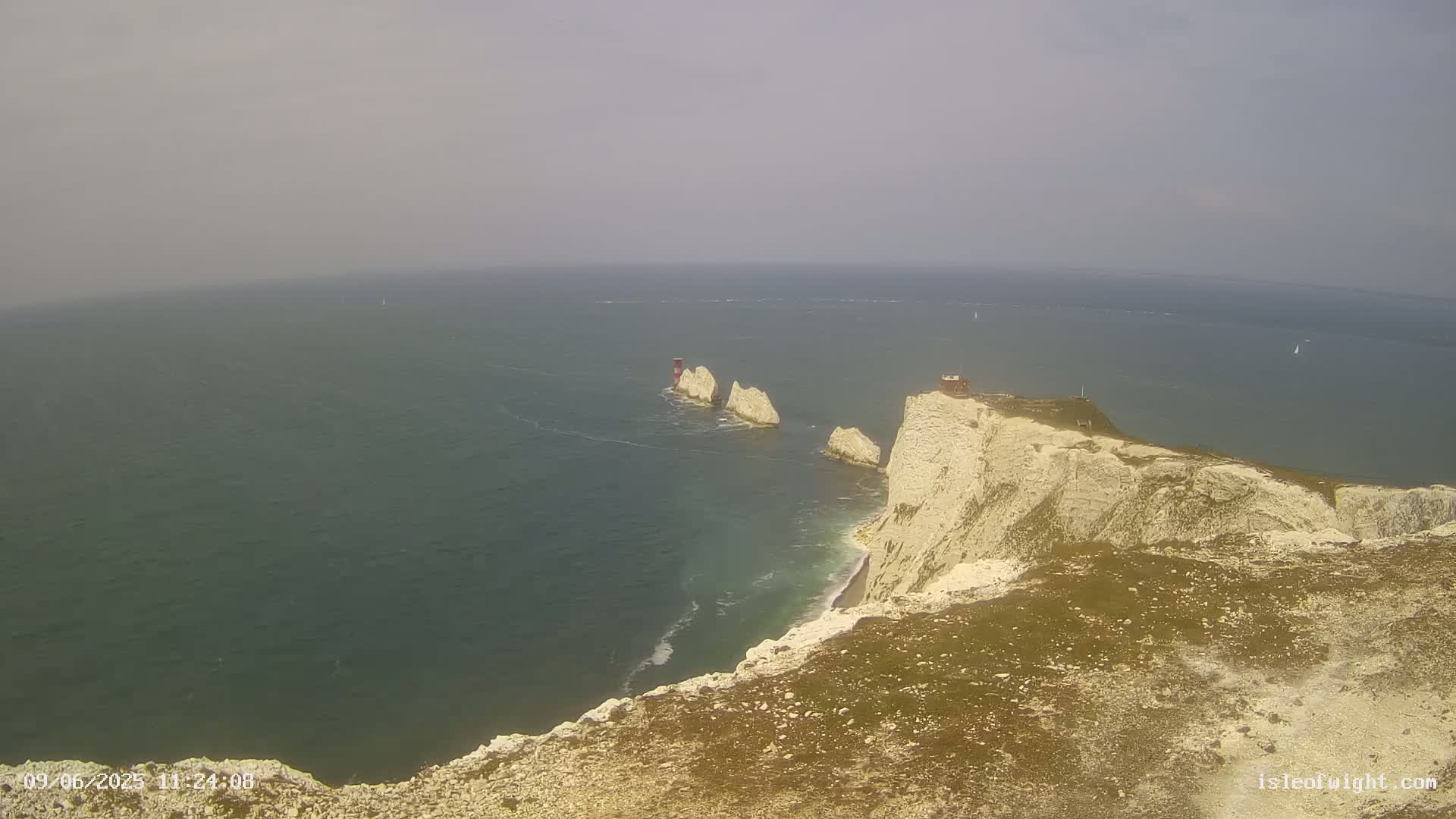 A hazy, overcast day reveals a high-angle view of white cliffs meeting a dark-blue sea with small waves, and three white rock formations with small structures jutting out from the water.