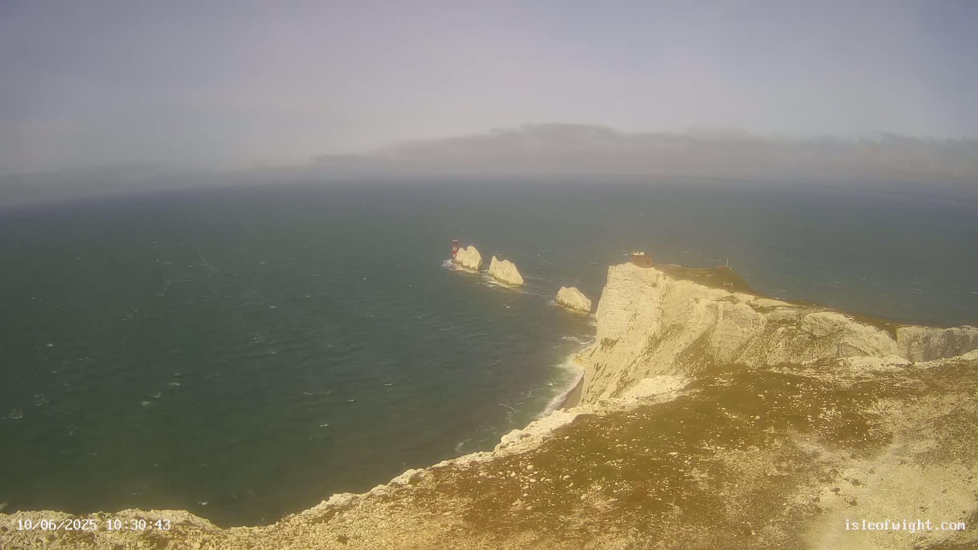 A hazy, windy day reveals a rocky coastline with a lighthouse and several stacks jutting out into a dark green sea.