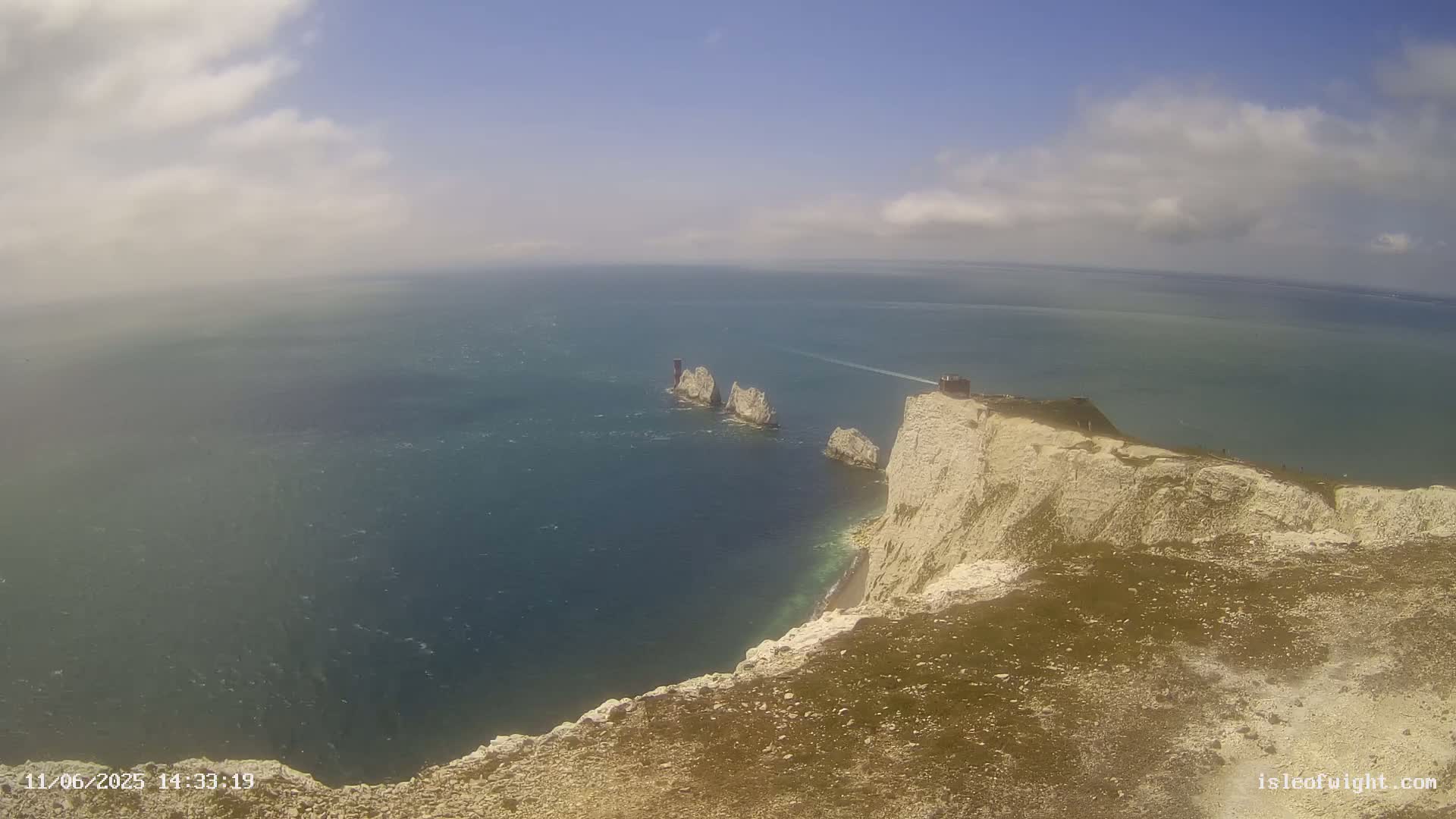 A high-angle view shows a partly cloudy, sunny day overlooking a white cliff overlooking the ocean with a small group of rocky islands in the distance.