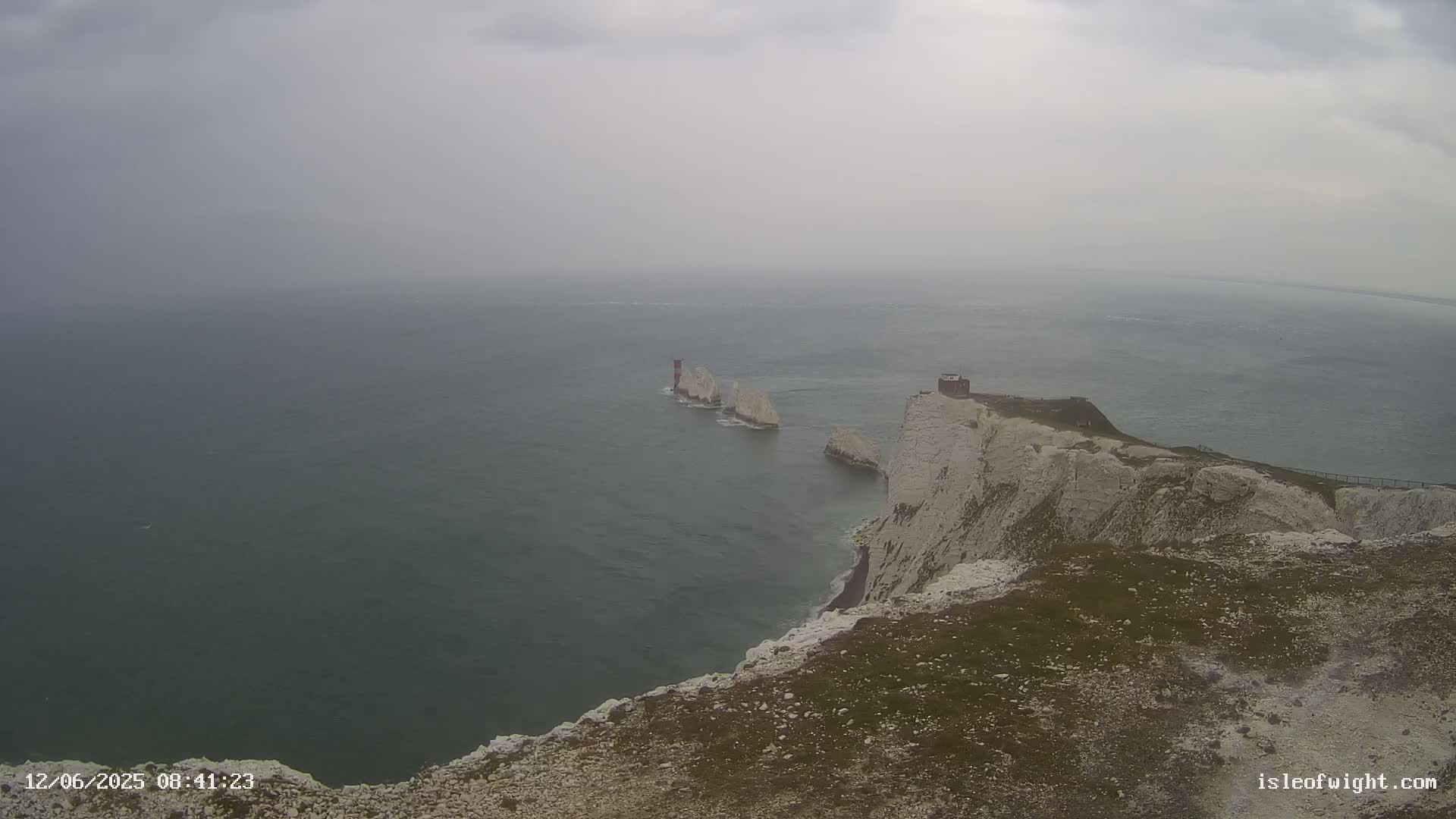 White cliffs overlooking a hazy, grey sea with several smaller white rock formations and a small building visible in the distance.