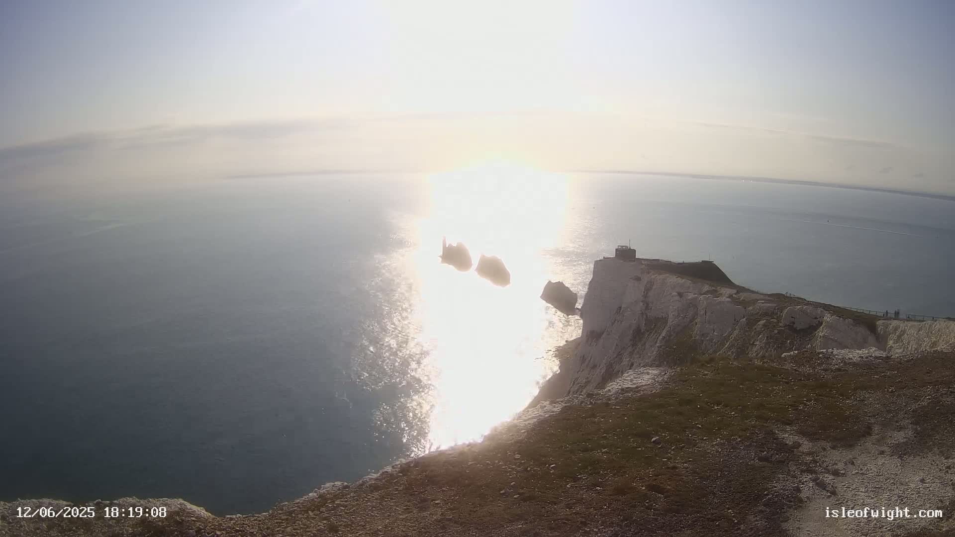White cliffs overlooking a calm sea with two small islands and a bright sun reflecting on the water.