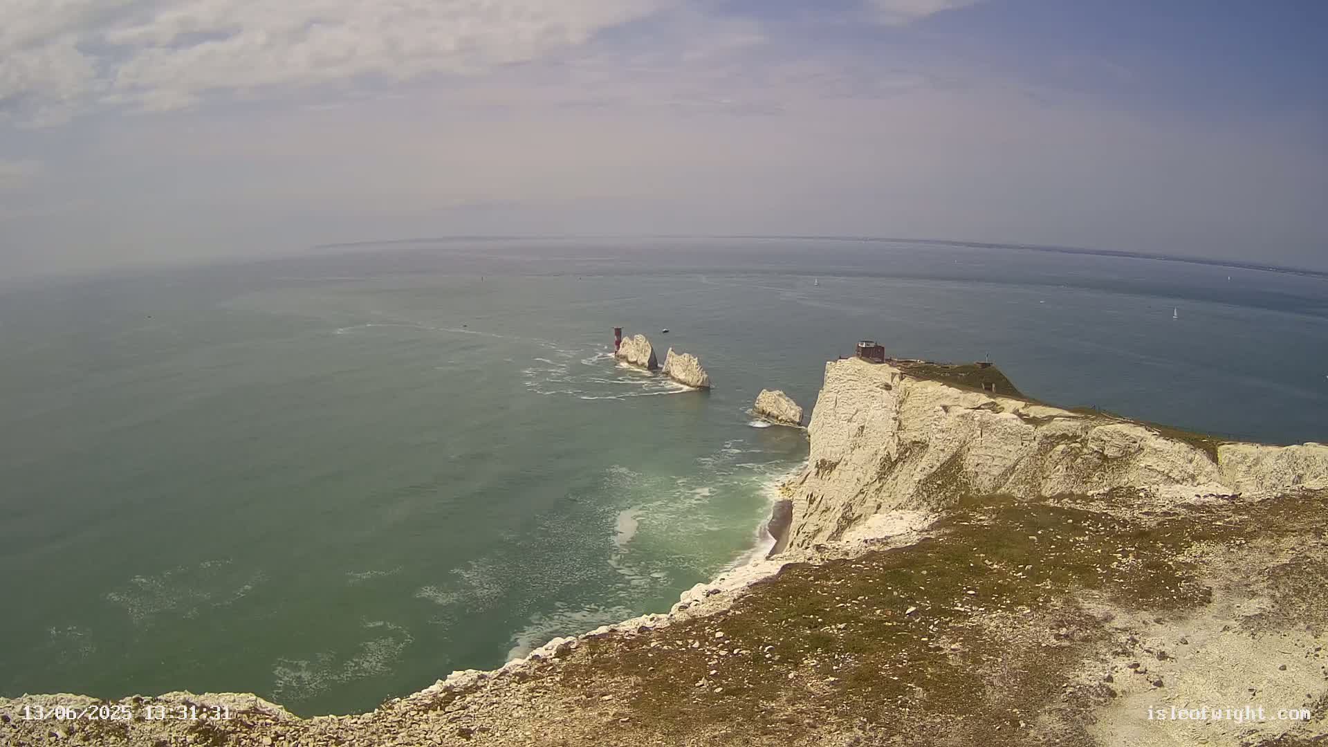 A high-angle view shows white cliffs meeting a calm, greenish-blue sea with several small, distant islands and a hazy sky.