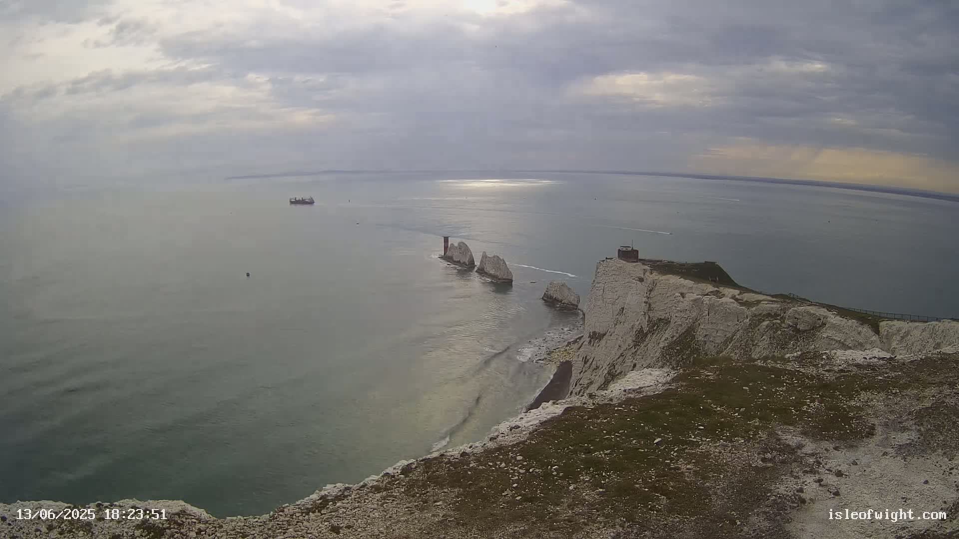 A view from a clifftop shows white chalk cliffs meeting a calm sea where several rock formations jut out from the water under an overcast sky.